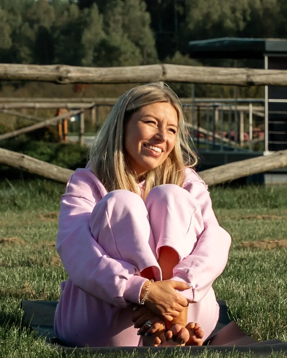 Ewa Wardęga in a pink tracksuit sits on the grass and smiles, with a wooden fence and a forest behind her