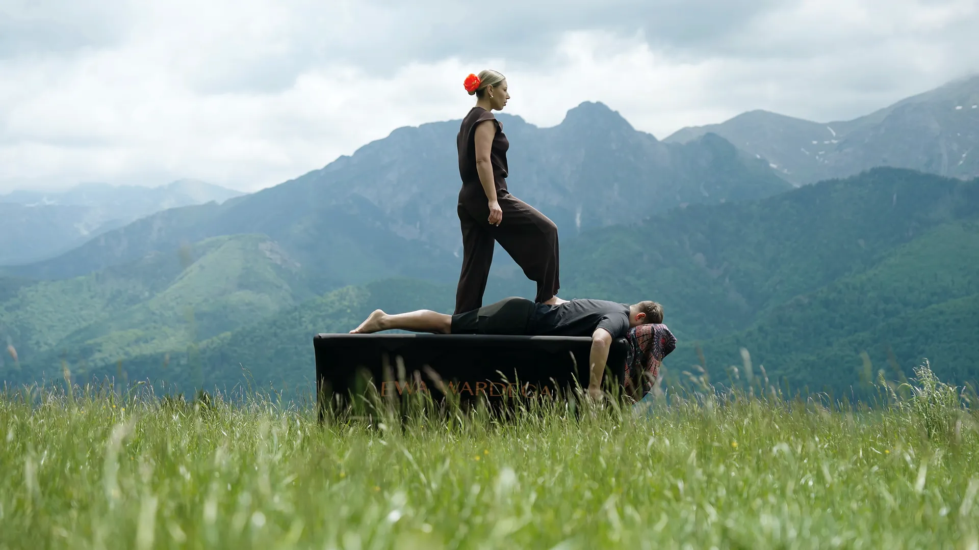 Ewa Wardęga stoi na stole do masażu nad leżącym klientem, stół stoi na łące, w tle Tatry